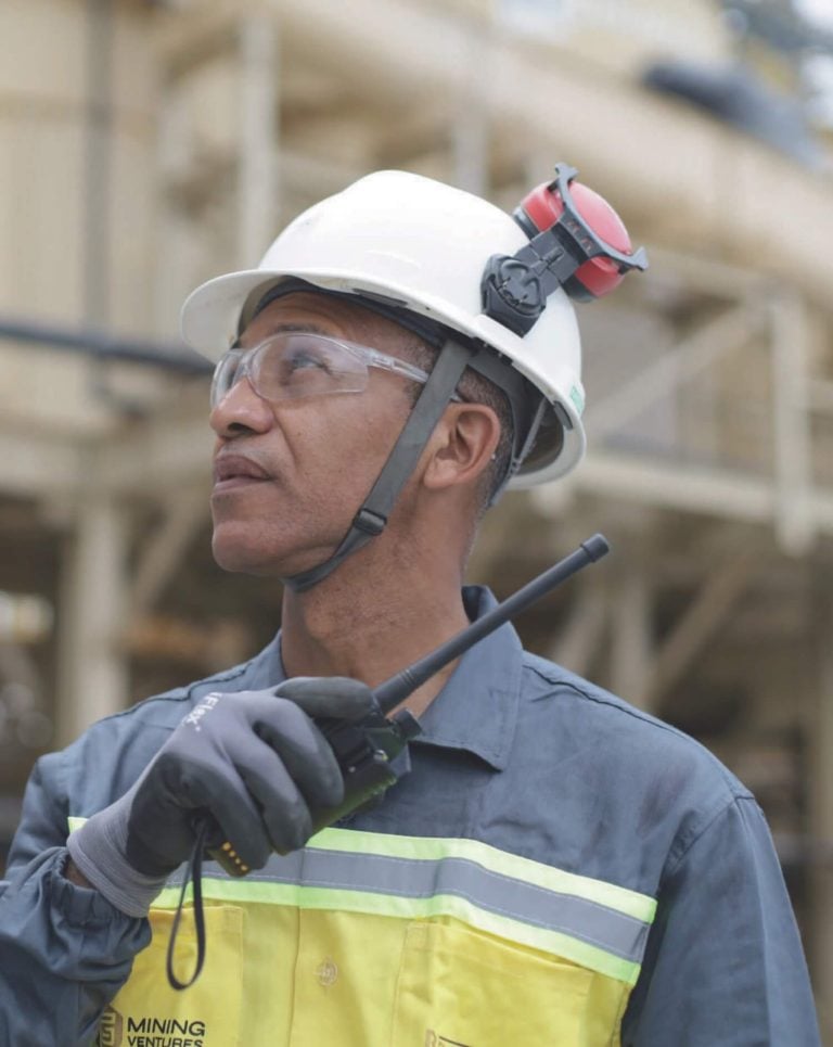 G Mining Ventures site operator wearing a white hard hat, safety glasses, and high-visibility workwear holding a two-way radio at a processing plant facility.