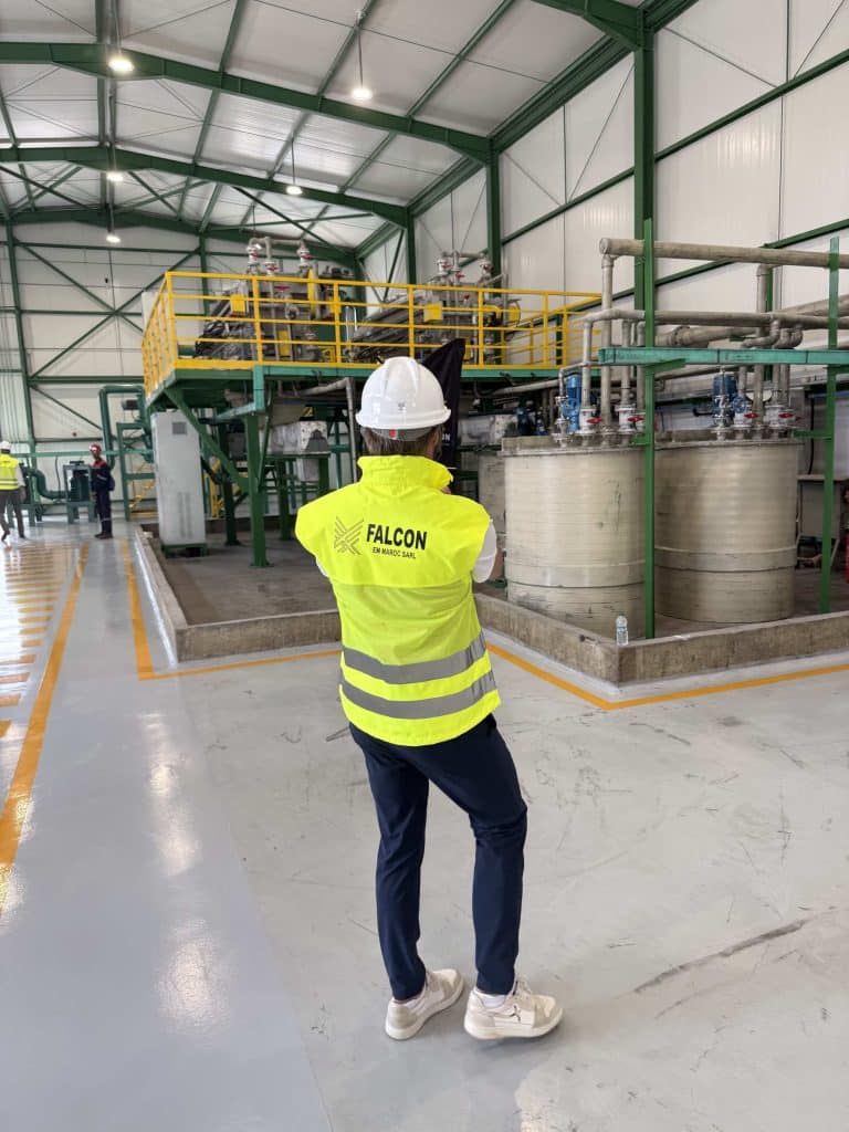 View of a worker wearing a white hard hat and high-visibility vest branded with the Falcon logo observing processing equipment inside a chemical refining plant.