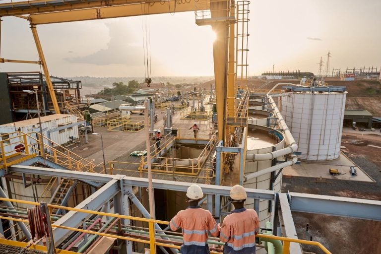 Two Endeavour Mining workers wearing hard hats and high-visibility shirts overlooking a gold processing plant structure in West Africa.