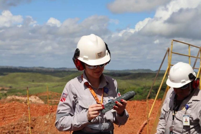 An Endeavour Mining geologist examining a drill core sample at a West Africa gold exploration site wearing safety helmet and ear protection.